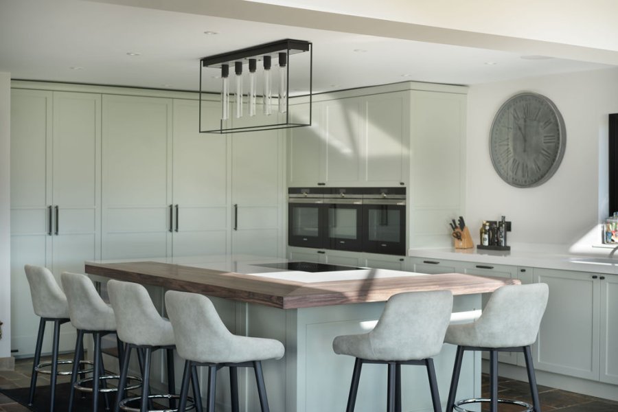 Modern kitchen with light grey cabinetry, a large island with a wooden countertop, and grey upholstered bar stools, illuminated by a contemporary pendant light.