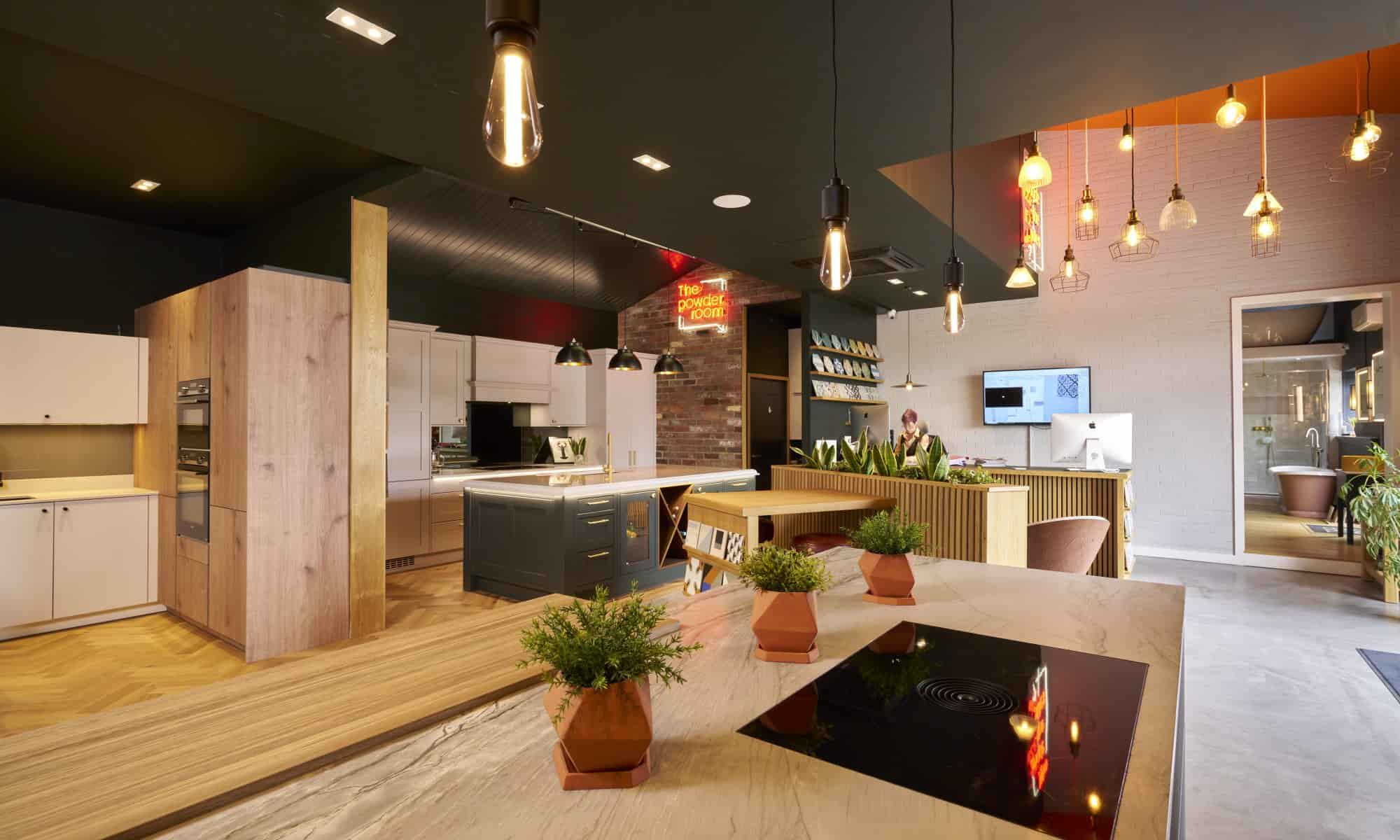 Contemporary kitchen showroom with multiple kitchen setups, featuring various cabinetry styles, a central island with plants, and modern pendant lighting.