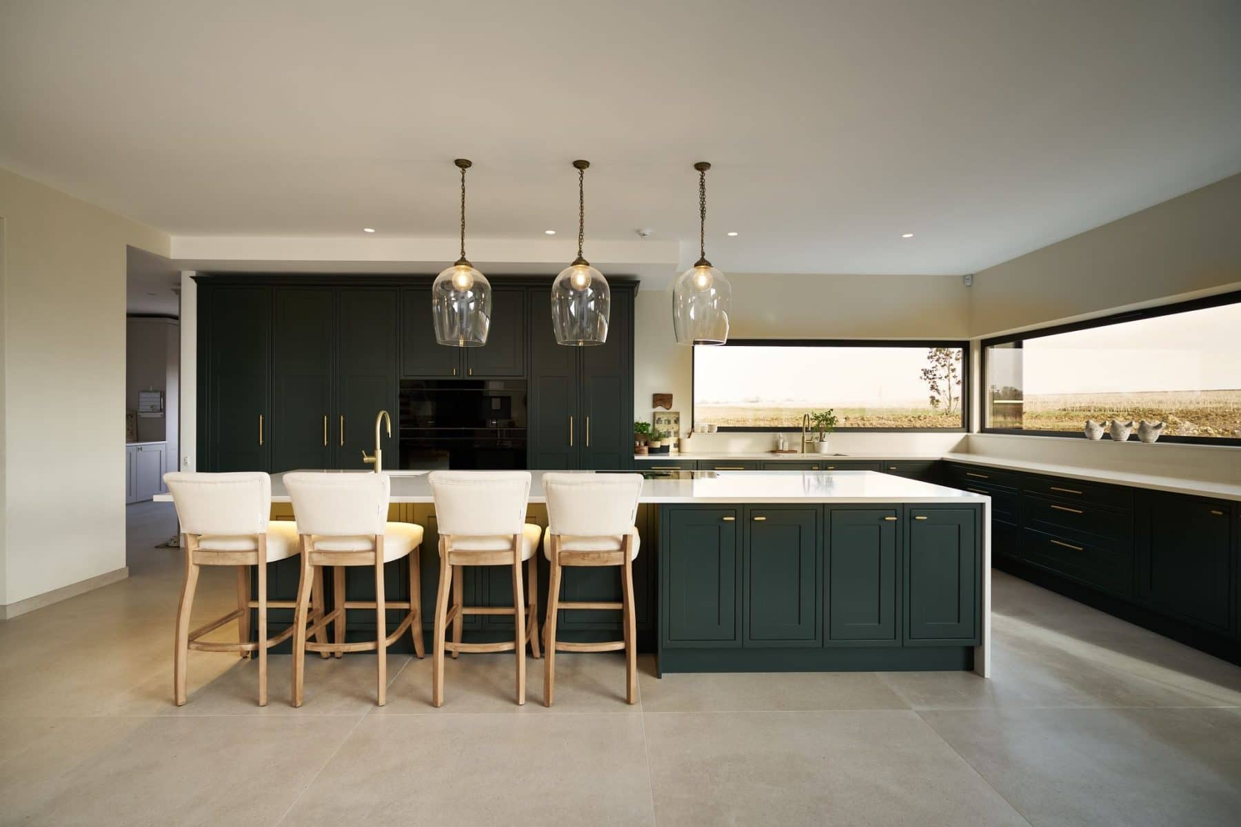 A modern kitchen with dark green shaker cabinetry, a large island with white countertops and light wooden bar stools, illuminated by three pendant lights, and featuring large windows with scenic views.