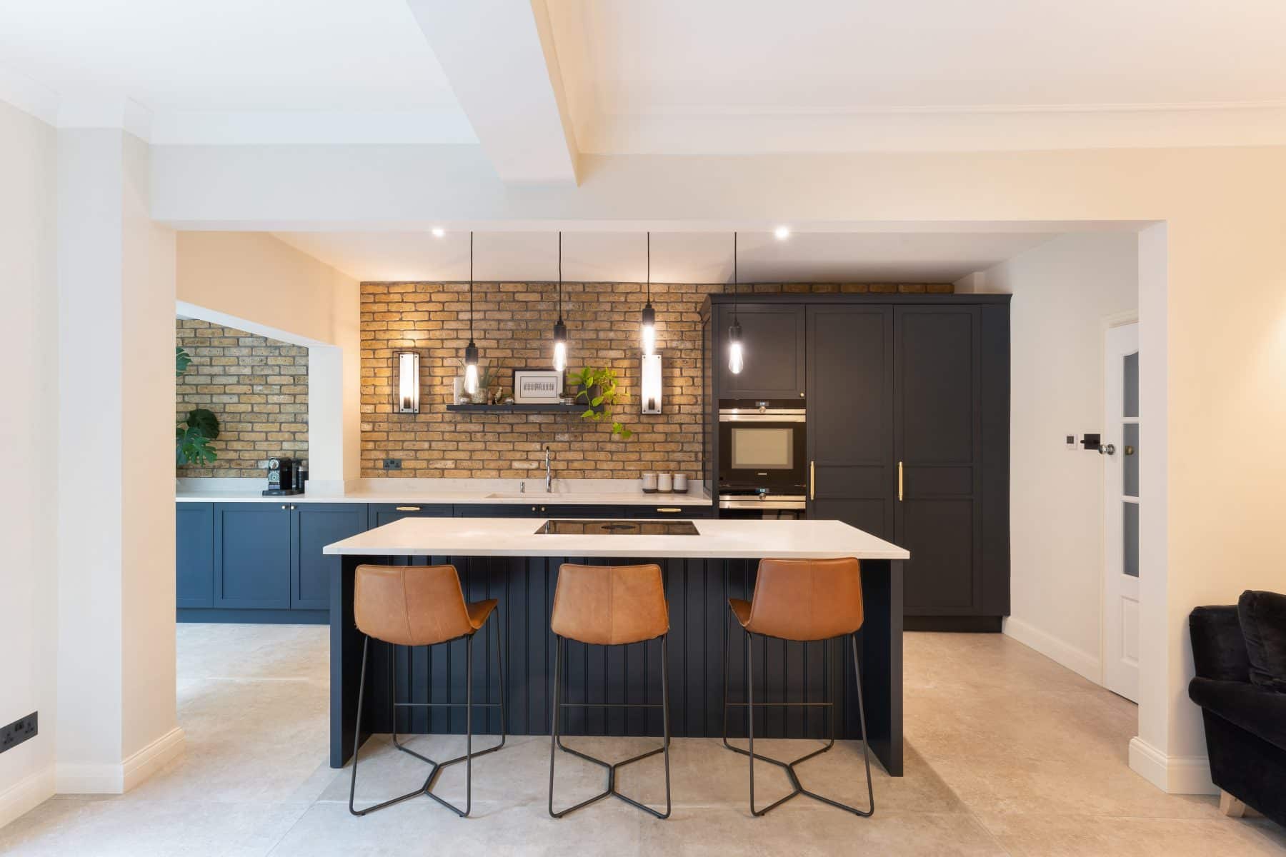 Modern kitchen with a black island featuring a built-in hob with white countertops and three brown, leather bar stools, and exposed brick walls, illuminated by black pendant lights.
