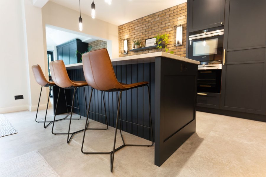 A close-up view of a modern kitchen island with white countertops, black cabinetry, and brown leather bar stools, set against an exposed brick wall.