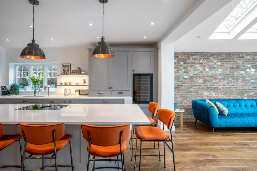A modern kitchen with a large white island, orange bar stools, light grey cabinetry, and a blue sofa in the adjacent living area, featuring exposed brick walls.