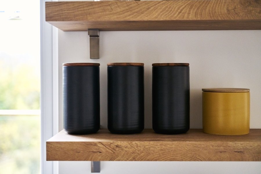 Three black jars with wooden lids and one yellow jar on a wooden shelf, against a light background.