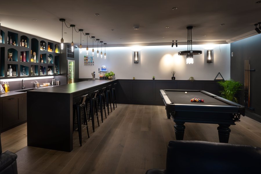 A modern home bar with dark cabinetry, a long counter with bar stools, and a pool table, illuminated by pendant lights.