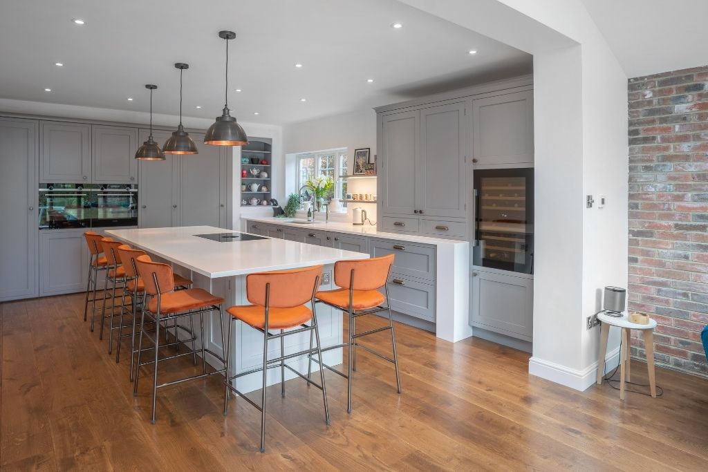 A contemporary kitchen featuring a large white island with orange chairs, grey cabinetry, and wooden flooring.